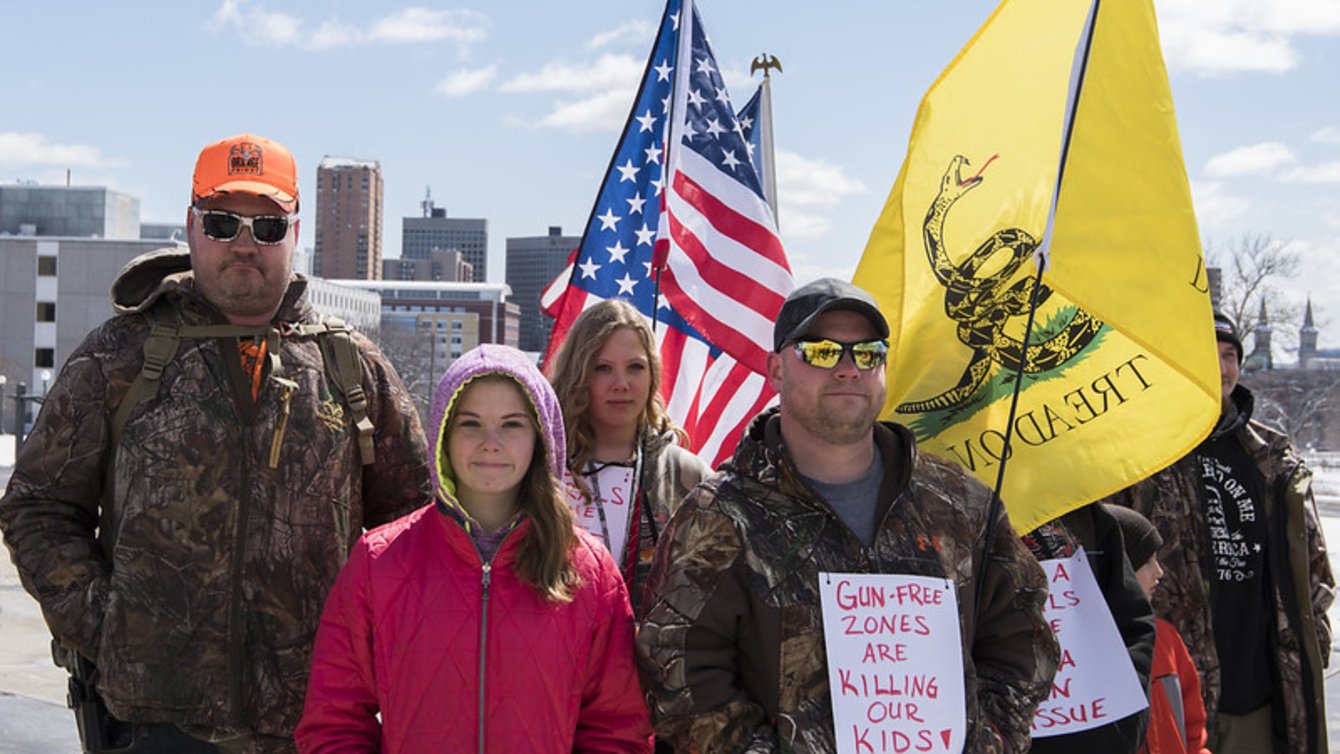 Small group poses for camera with US and Gadsden flags. In front, a child in pink jacket stands beside man wearing a brown camo jacket with sign pinned on it. Sign reads: "GUN FREE ZONES ARE KILLING OUR KIDS"