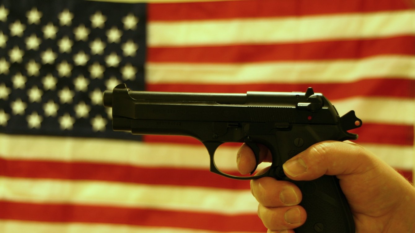 A hand gripping a handgun in front of an American flag backdrop