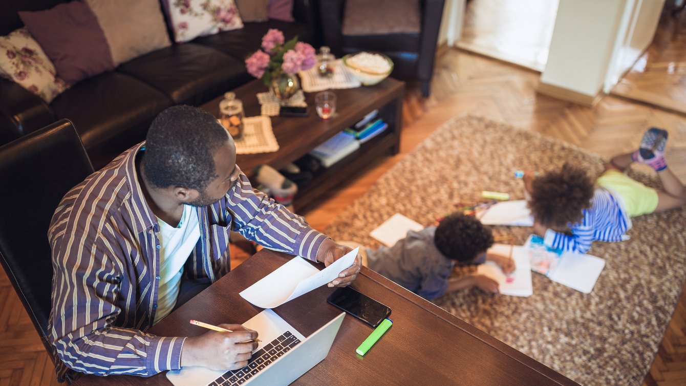 Adult man works on laptop and looks after two children reading nearby in room.