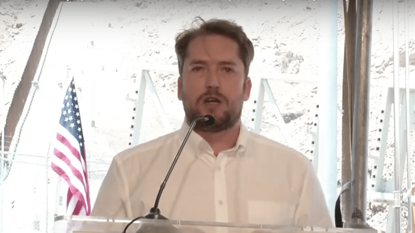 Darren Beattie delivers speech in NM with U.S.-Mexico border wall construction behind him. 