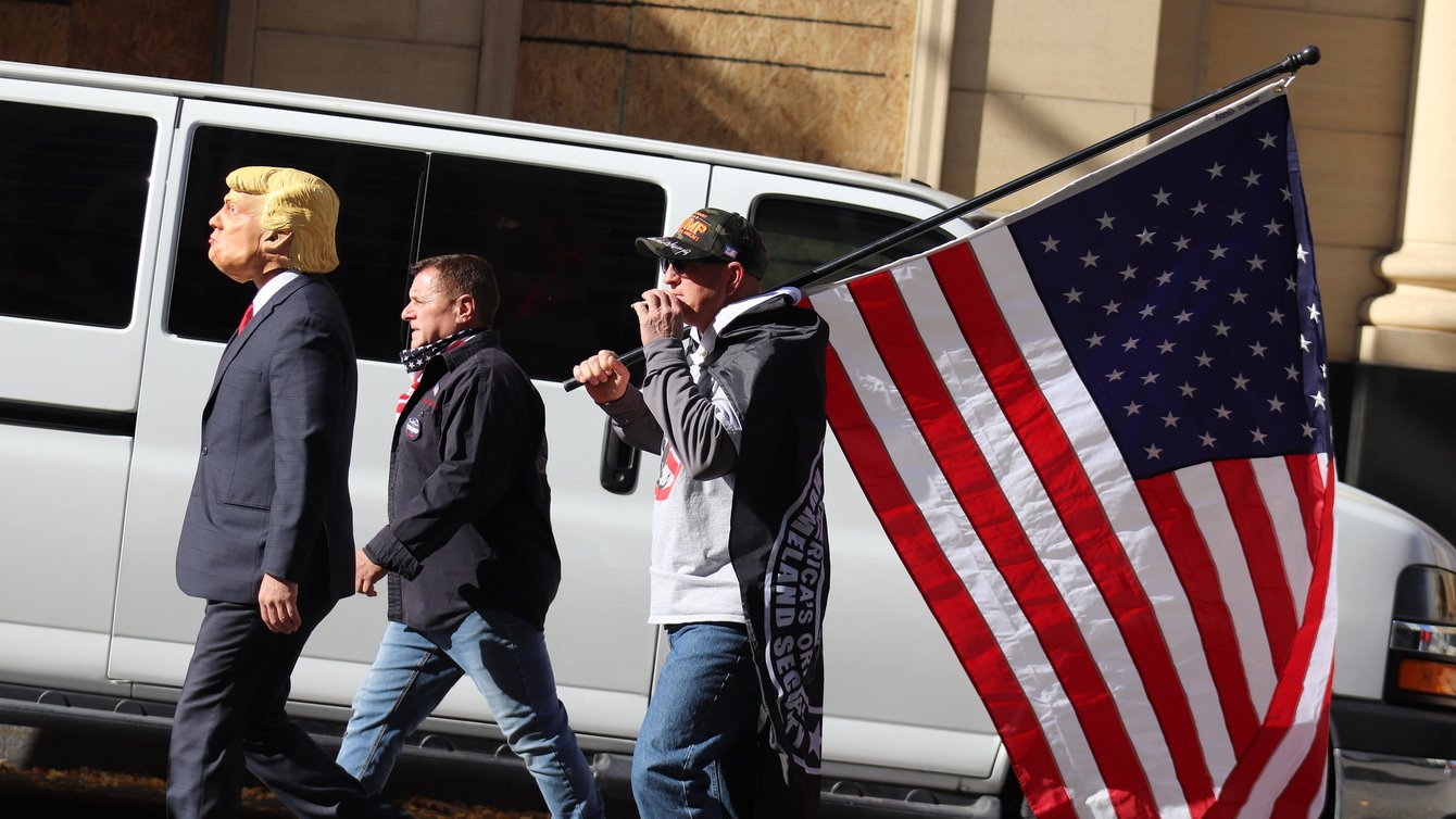 Three men walking; one wears a Trump mask and one carries an American flag