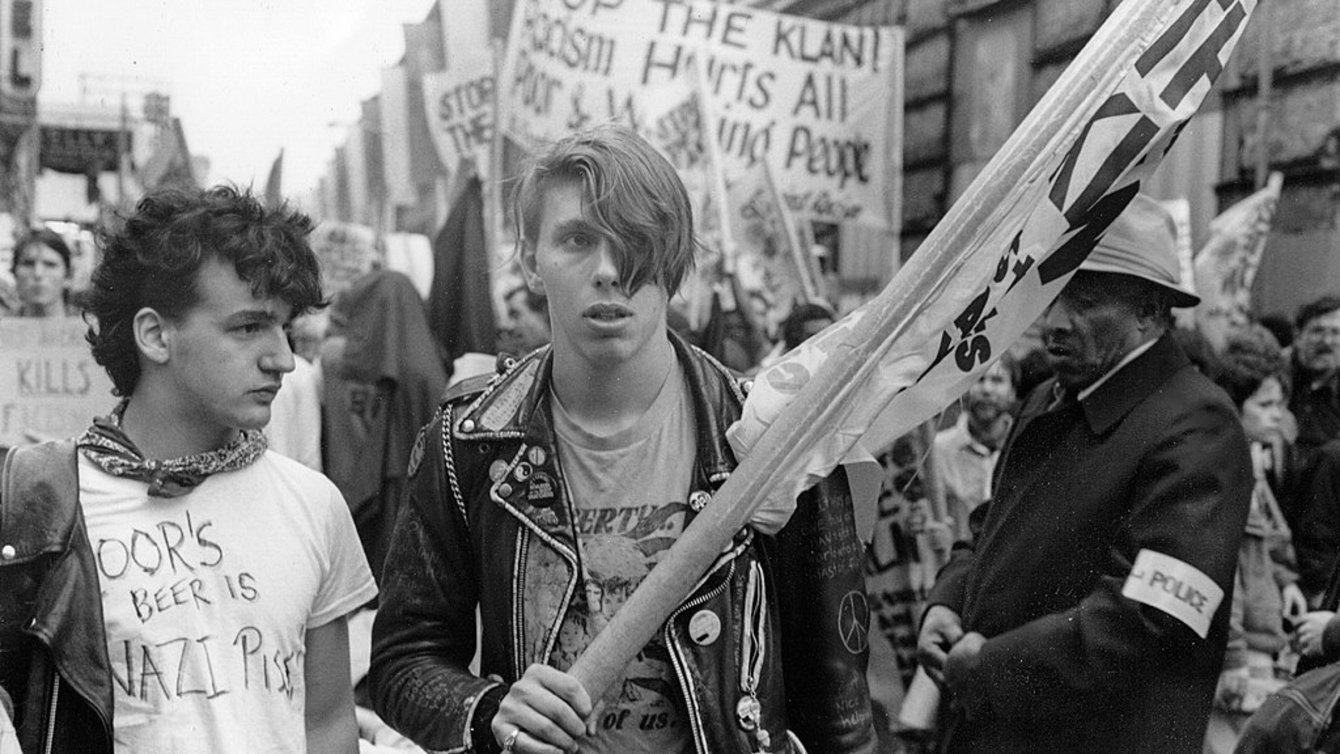 Two people at a protest. One is holding a flag, wearing a leather jacket. The other man is wearing a white t-shirt with anti-nazi writing. 