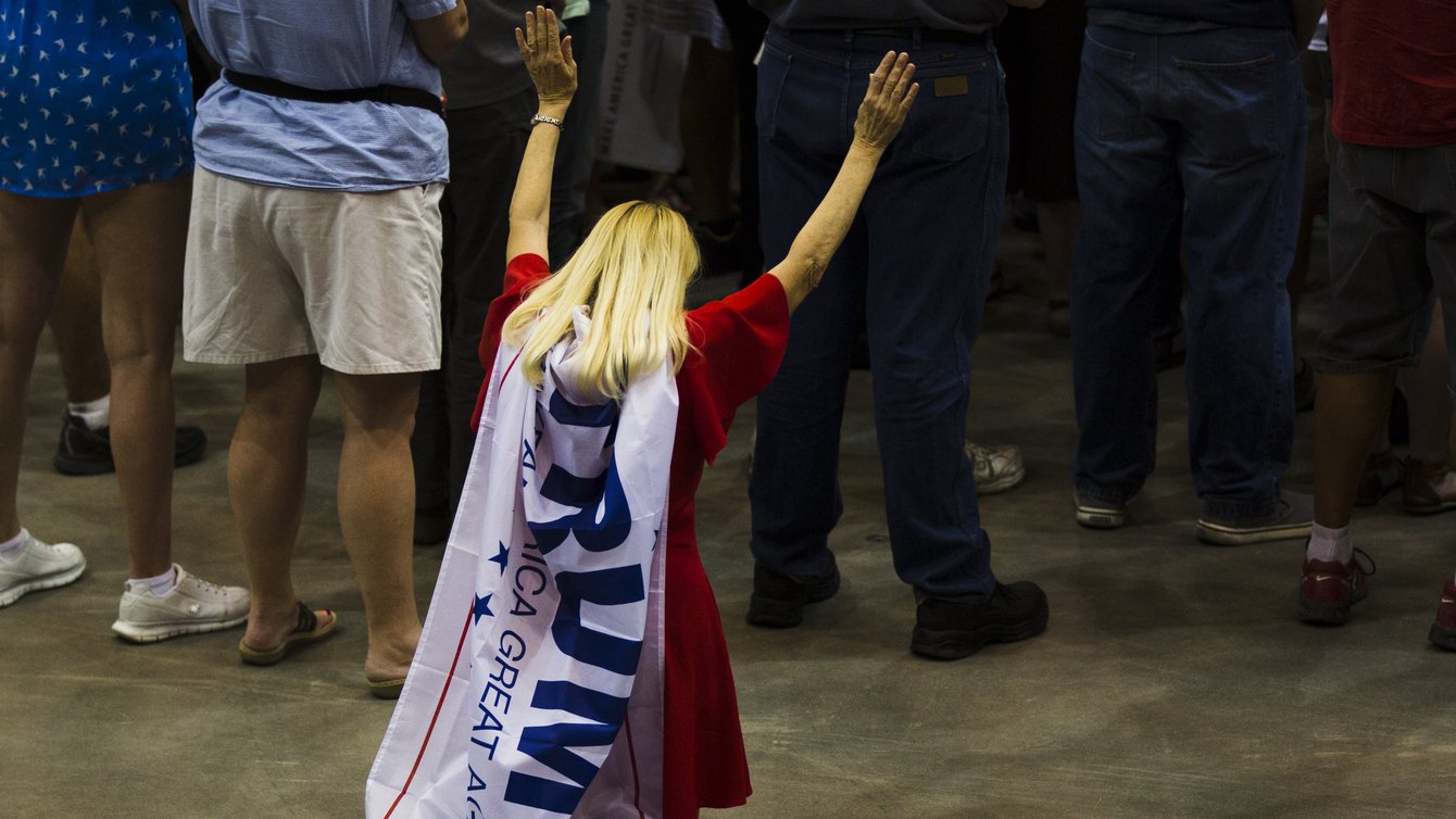 A white woman with blond hair, kneeling with her arms in the air, a Trump flag tied around her neck and hanging down her back
