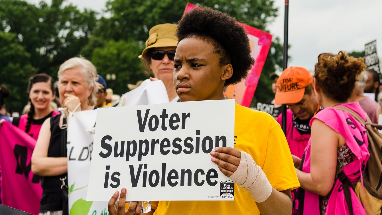 A Black person in a yellow T-shirt holding a sign that says "voter suppression is violence"