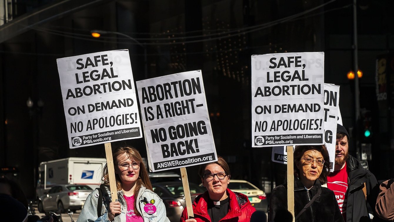 Three women at a prochoice rally holding signs that say "Safe, legal abortion on demand. No apologies!" and "Abortion is a right. No Going Back."