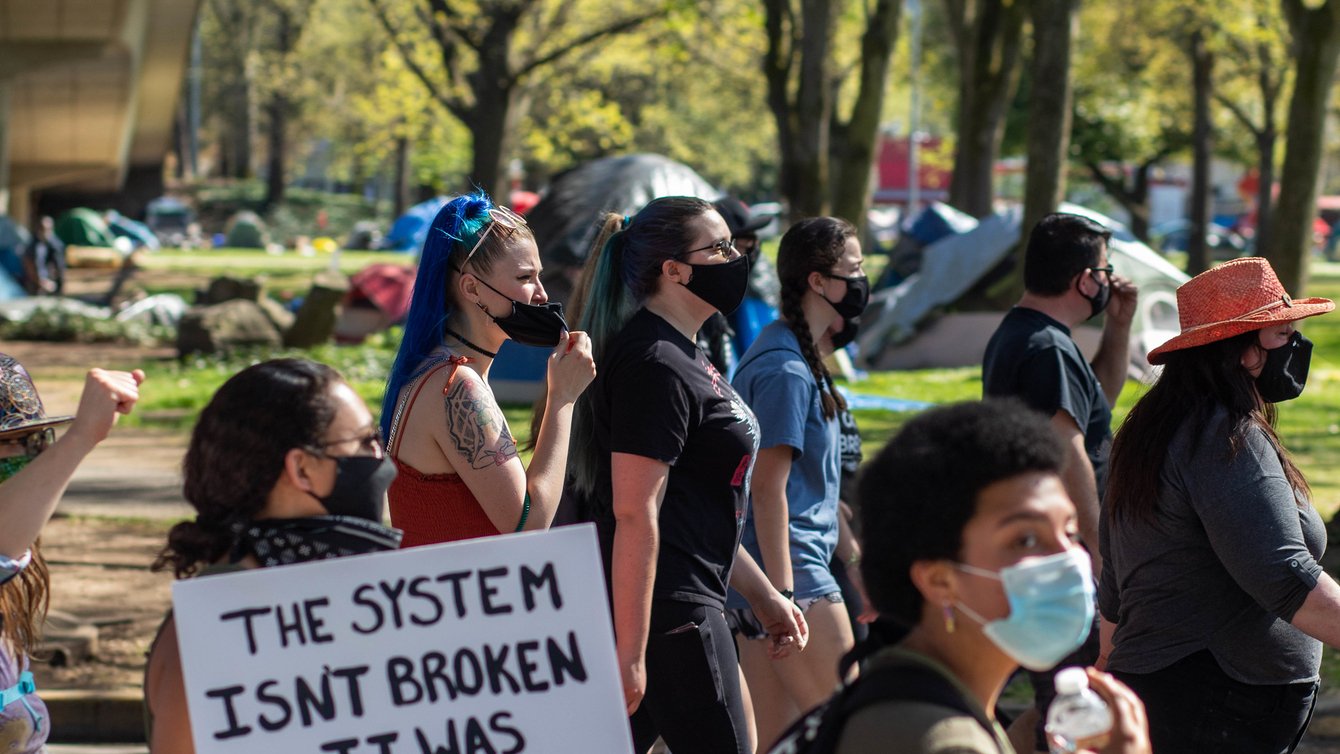 Protesters wearing masks. One protester holding a sign that says "The system isn't broken it was built this way."