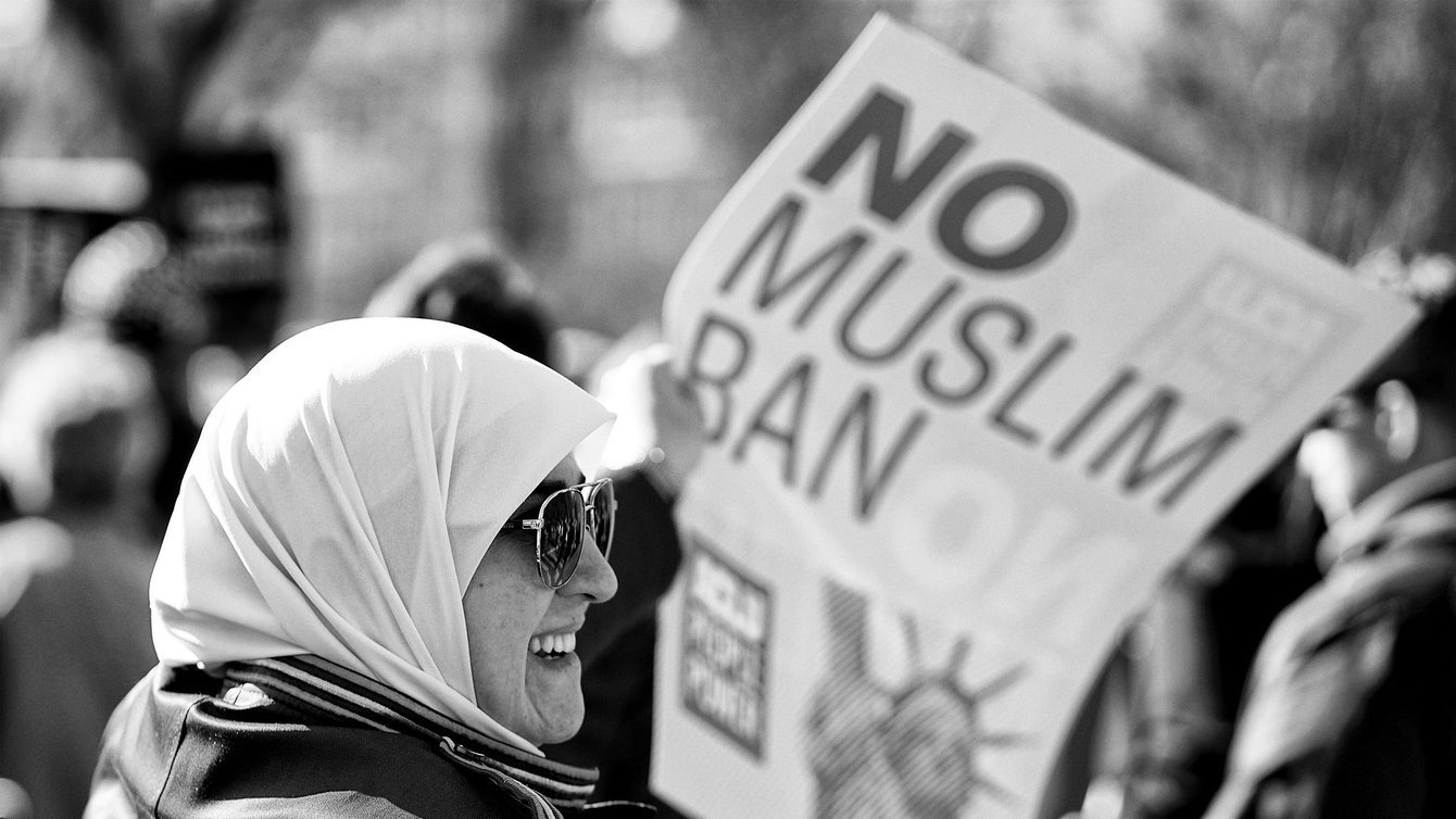 A woman in a hijab smiling, holding a poster that says No Muslim Ban
