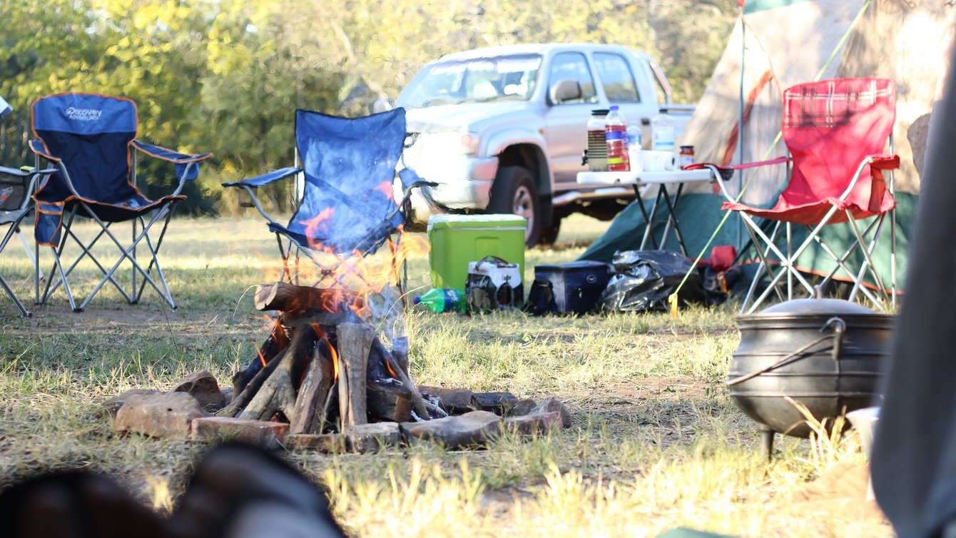 Stock photo of a camp with fire, camping chairs, the side of a tent, and a silver pickup truck in the background