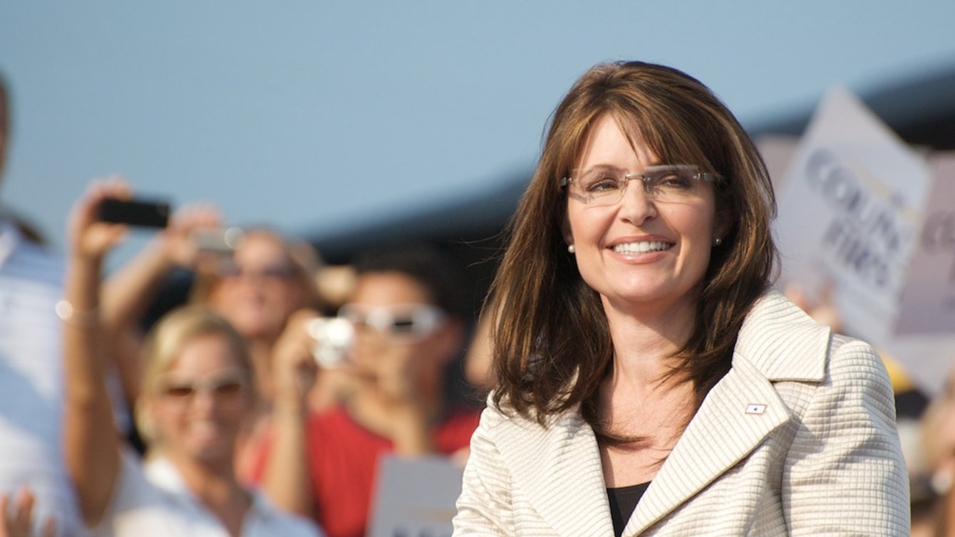 Sarah Palin in a white jacket, in front of a crowd, smiling.