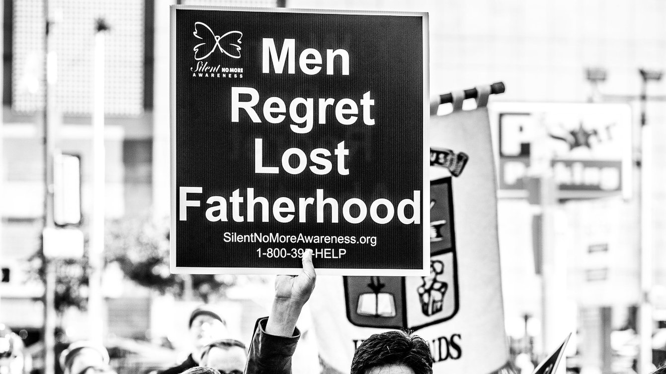 A man wearing sunglasses holding a sign that says "Men Regret Lost Fatherhood"