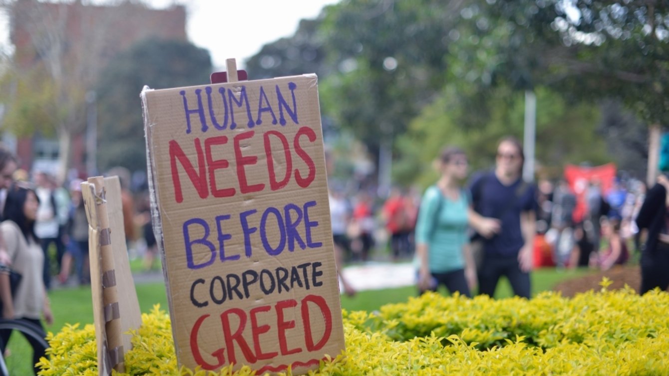 A cardboard sign that says Human Needs Before Corporate Greed