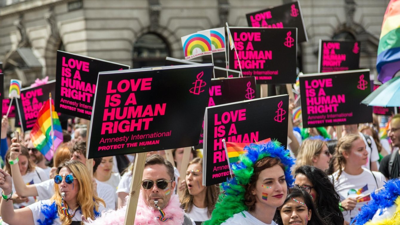 People in a protest wearing rainbow wigs, holding signs that say "love is a human right"
