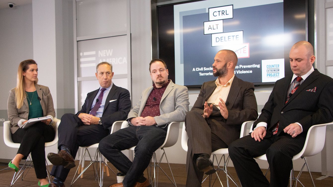 A white woman and four men sitting in chairs in straight line. Behind them a board says "Ctrl Alt Delete Hate".
