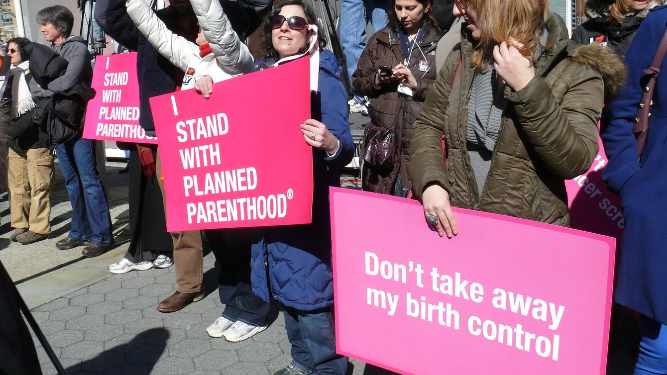 Protesters holding banners. One says I Stand with Planned Parenthood, another one says "Don't take away my birth control"