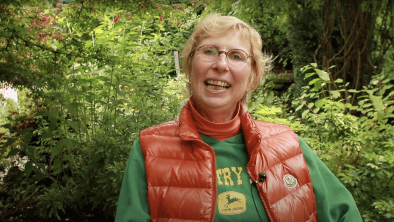 A White woman with short blond hair wearing a green t-shirt and orange sleeveless jacket sitting in front of some bushes.