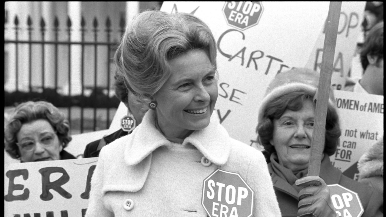 A White woman in a heavy coat wearing a "Stop ERA" badge smiles with other protesters in front of the White House fence. 