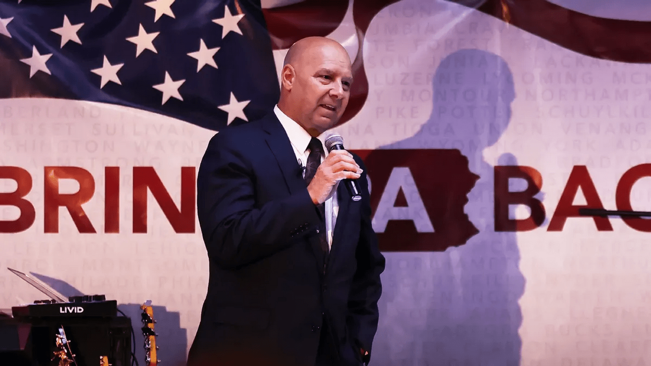 A bald man in a suit, standing in front of an American flag, talking into a mic.