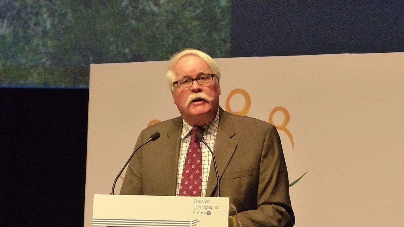 A man with white hair and a white mustache, speaking into a microphone.