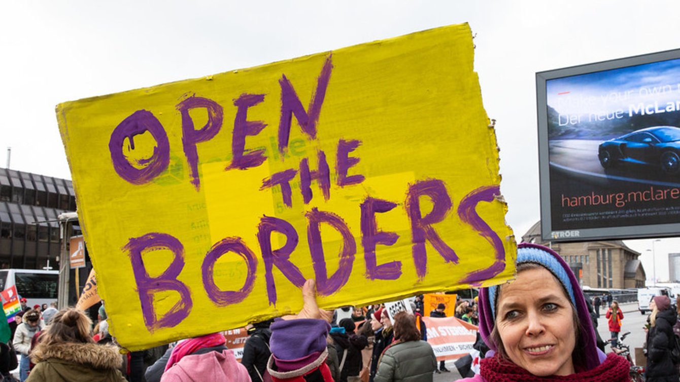 A woman with a scarf holding a sign that says "open the borders"