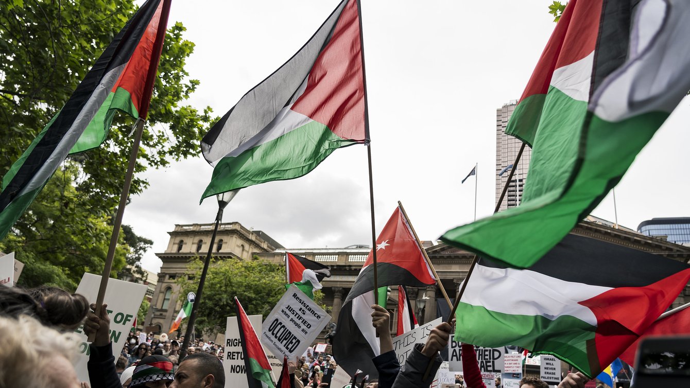 People protesting, waving red, white, and green Palestine flags in the air.