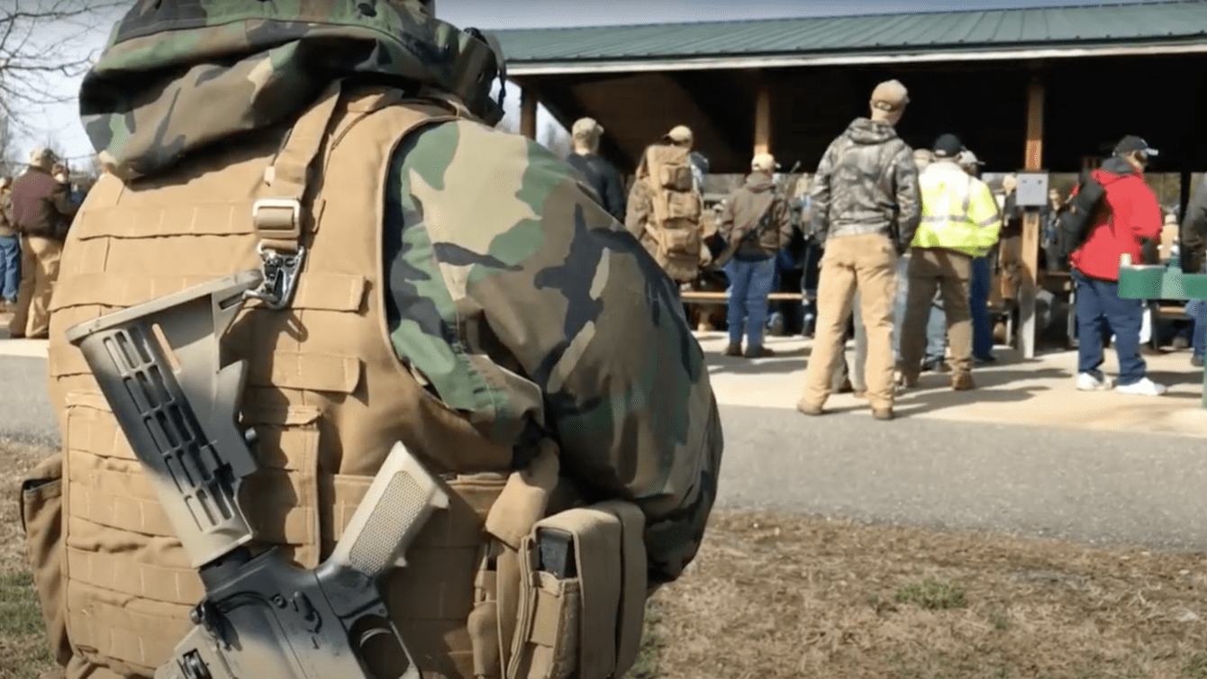 A man in camouflage in the foreground with a gun on his shoulder.