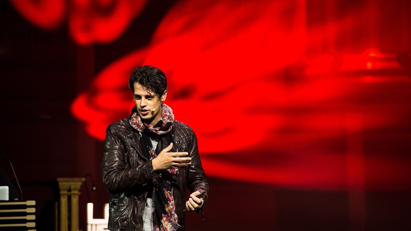 A man in a black jacket speaks in front of a red blurred background 