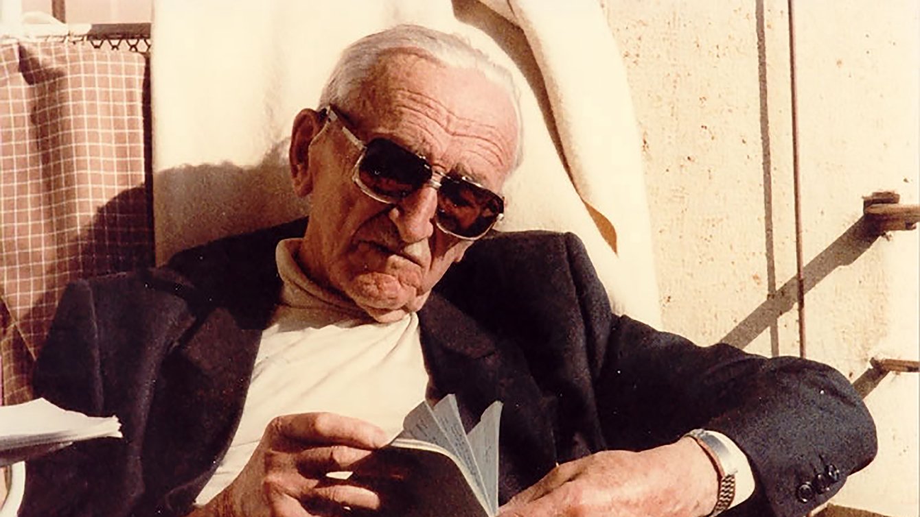 old man with sunglasses and white hair reads a book in the sun