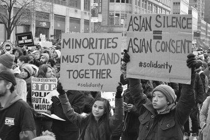 a black and white image of protestors. They're holding signs that read "Minorities must stand together" and "Asian Silence = Asian Consent"