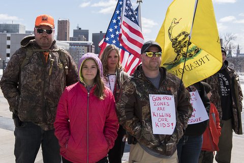 Small group poses for camera with US and Gadsden flags. In front, a child in pink jacket stands beside man wearing a brown camo jacket with sign pinned on it. Sign reads: "GUN FREE ZONES ARE KILLING OUR KIDS"