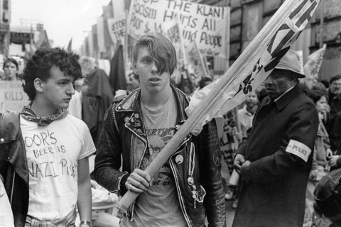 Two people at a protest. One is holding a flag, wearing a leather jacket. The other man is wearing a white t-shirt with anti-nazi writing. 