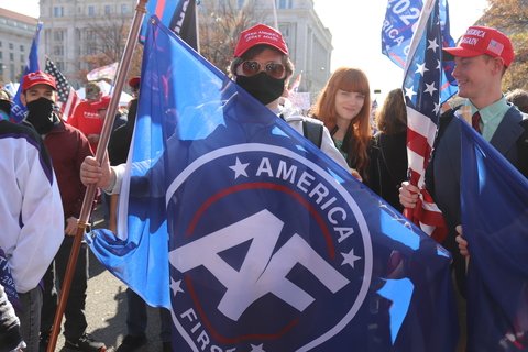 A man holding an "America First" flag, wearing a red MAGA hat, and a black face mask in a crowd of other right-wingers.