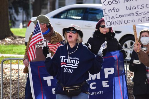 A White woman with blond hair, wearing a hat. She has a Trump 2020 sweatshirt on, and is holding an Amerian Flag and a water bottle. Her mouth is open like she's shouting something.