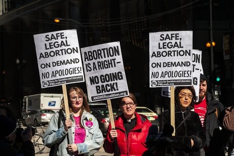 Three women at a prochoice rally holding signs that say "Safe, legal abortion on demand. No apologies!" and "Abortion is a right. No Going Back."