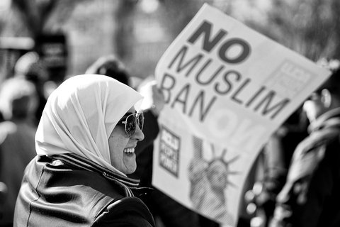 A woman in a hijab smiling, holding a poster that says No Muslim Ban