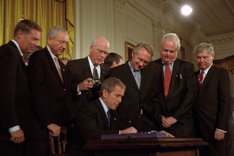 Presdient George Bush signing something, with men standing around him.