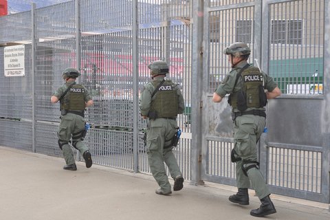 Three men in army training suits running along a large metal fence
