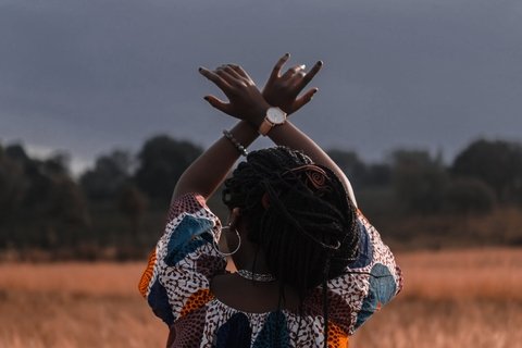 Black woman in field facing away from camera with arms in the air crossed at the wrist. 
