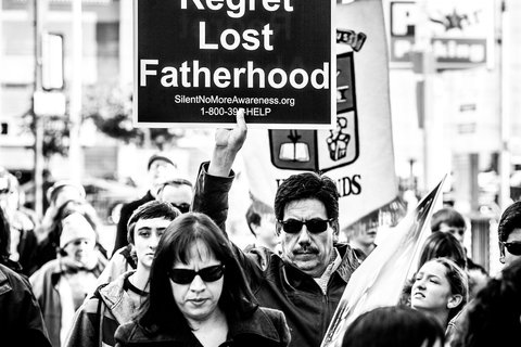 A man wearing sunglasses holding a sign that says "Men Regret Lost Fatherhood"