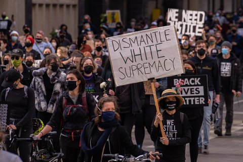 A person wearing a mask at a protest holding a sign that reads "Dismantle White Supremacy"