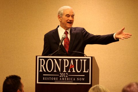 A man in a suit talking into a mic and pointing to his left. A sign on the podium says "Ron Paul, 2012, Restore America Now"