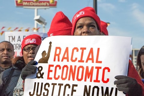 Woman in hat and gloves holds sign reading "Racial Economic Justice Now."