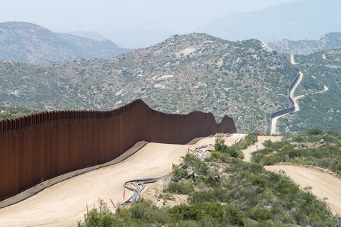 a brown fence following around a dirt road that fades into green mountains.