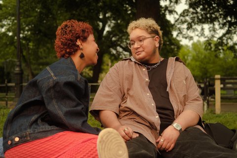 Two trans people in a park, looking at each other, laughing.