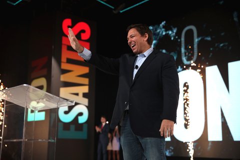 White man on stage with hand raised smiling, you can see the letters "ESANTIS" projected behind him.