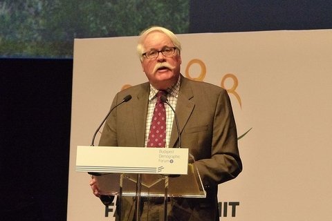 A man with white hair and a white mustache, speaking into a microphone.