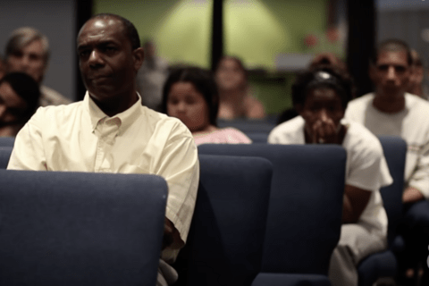A black man in the foreground sitting in a grey chair, people behind him.
