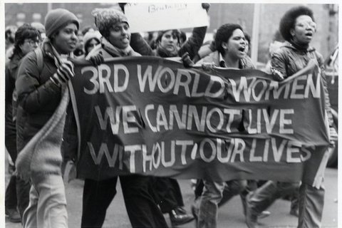 Four people walking holding a banner that says "3rd world women we cannot live without our lives"