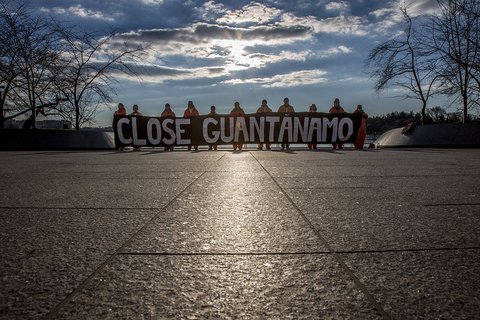 A row of people in orange jumpsuits holding a sign that says "close Guantanamo"