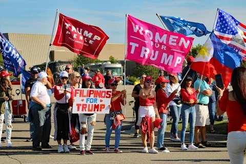A group of women in red hats hold flags reading "Women for Trump," "Filipinos for Trump," and "Trump pence"
