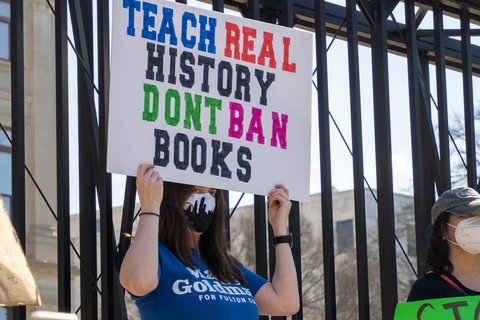 Woman in blue shirt holding a sign that reads "Teach real history don't ban books"